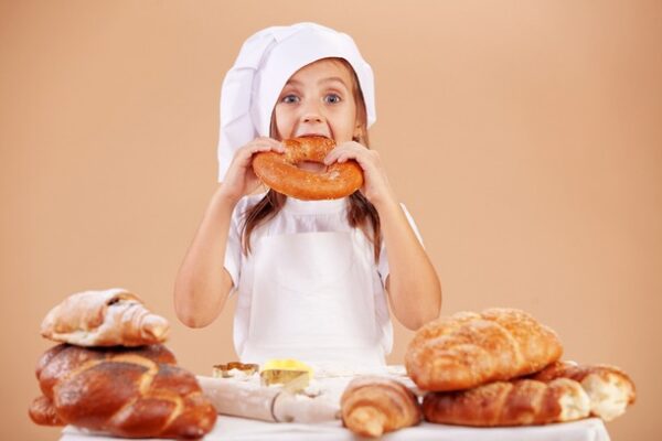 A small child eating a large pretzel and surrounded by bread
