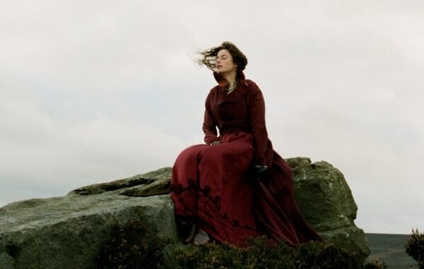 A woman wearing a long red dress sitting on a large rock on moorland